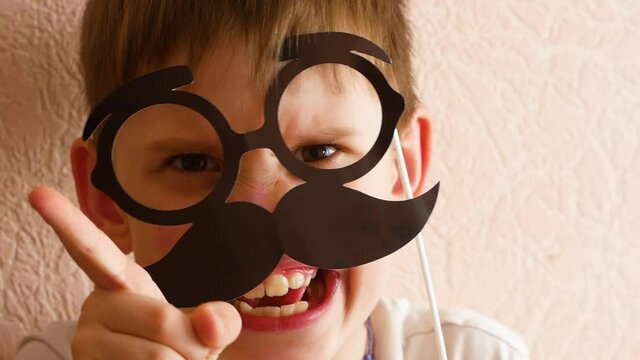 Happy Father's Day! A funny son in a dad mask with glasses and a mustache depicts a strict dad. Playful portrait of a Caucasian boy in mask glasses with a mustache. Selective focus, shallow depth of f