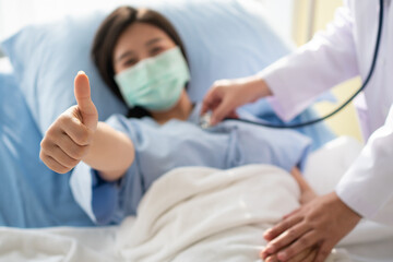 Happy Asian female patient wears a mask, lies on the bed, and raises her thumb up. When a doctor uses a stethoscope to listen to the lungs. Concept of believe in treatment And insurance coverage