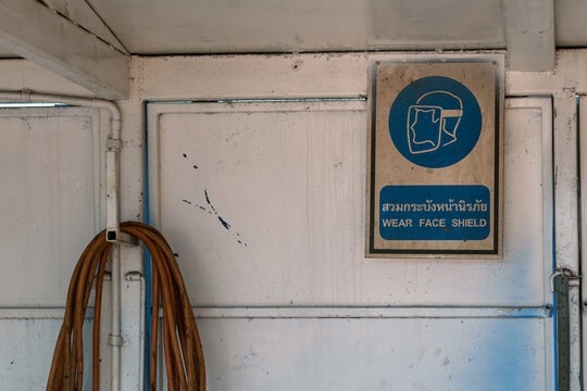 "Wear face shield" warning sign which is installed on welding workshop or garage wall. Industrial safety sign "Wear face shield" (Thai and English text) safety sign plate on the metal wall at welding 