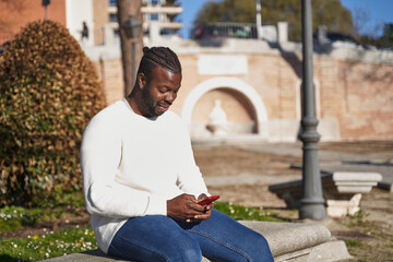 Obraz premium Young African American man sitting on a park bench sending a message. Latino man with anti-contagion mask. Concept of new normal. 