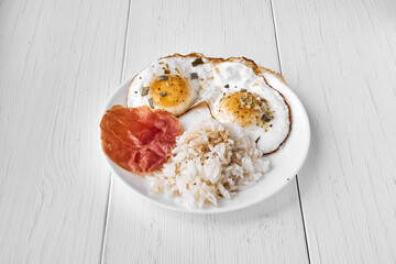 Two fried eggs sprinkled with dry green onions and a piece of bacon and rice topped with soy sauce for a side dish. Proteins, carbohydrates, fats are a balanced meal for lunch.