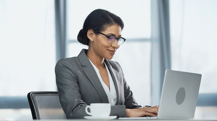 happy african american businesswoman using laptop near cup of coffee