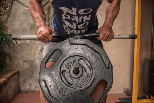 An anonymous man does T-bar rows at the gym. Wearing a tank top with the words No Pain, No Gain. Sweaty and muscular torso.