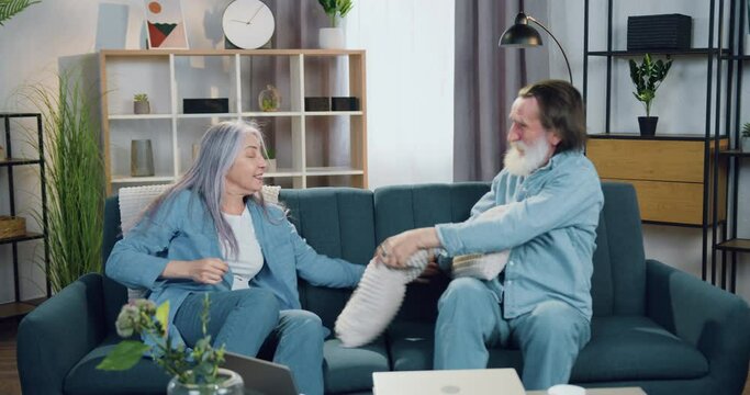 Close up of funny happy lovely old couple which sitting on comfortable sofa at home and fighting with pillows during joint leisure