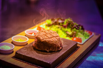 Grilled juicy beef steak smoking on hot stone served on wooden board with salad and sauces, colorful background