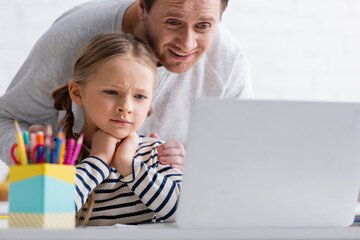 smiling man touching thoughtful daughter during online lesson at laptop on blurred foreground