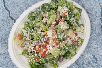 Overhead view of greek salad piled high on a paper plate with romaine lettuce, feta cheese, cucumber, and dressing to fill any appetite.