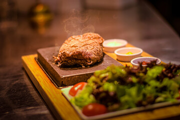 Grilled juicy beef steak smoking on hot stone served on wooden board with salad and sauces