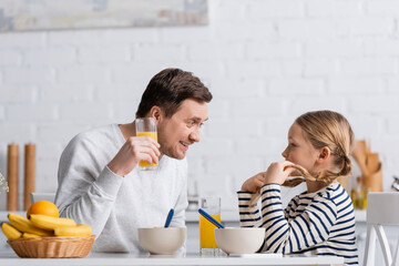 smiling man holding glass of orange juice while talking to daughter during breakfast