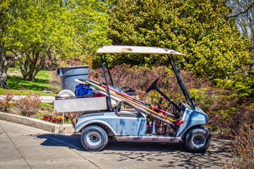Motorized golf cart being used as a garden maintenance vehicle parked on pavement in front of park.