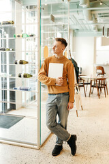 Young smiling business man freelancer in casual yellow sweater and jeans working remotely using laptop and phone sitting in cafe, selective focus