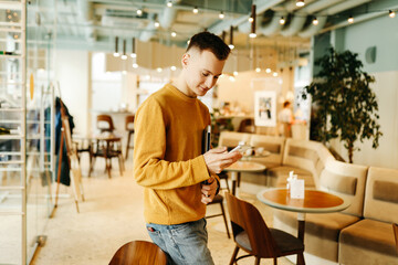 Young smiling business man freelancer in casual yellow sweater and jeans working remotely using laptop and phone sitting in cafe, selective focus