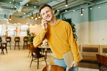 Young smiling business man freelancer in casual yellow sweater and jeans working remotely using laptop and phone sitting in cafe, selective focus