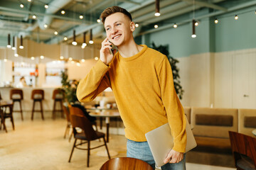 Young smiling business man freelancer in casual yellow sweater and jeans working remotely using laptop and phone sitting in cafe, selective focus