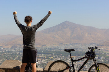 Young man with arms up next to bike with views of Santiago city on sunny day. Triumphant cyclist on...