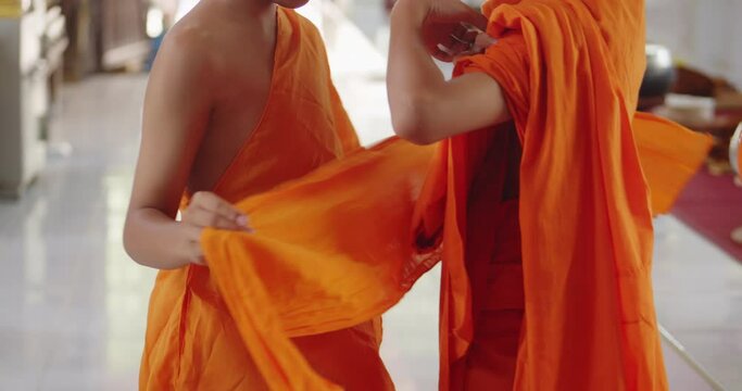 Slow motion scene of the Buddhist novice who has just passed ordination in the summer during school holidays is helping an other novice to dress in a yellow-orange robe.
