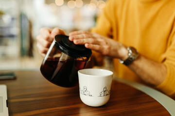 A smiling barista man in a bright yellow cozy sweater holds a teapot with his hands and pours tea into a mug in the indoor cafe