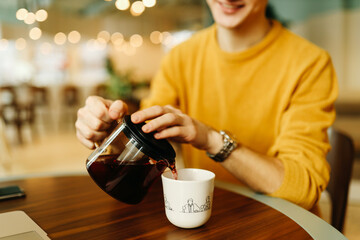 A smiling barista man in a bright yellow cozy sweater holds a teapot with his hands and pours tea into a mug in the indoor cafe