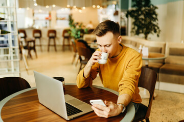 Young smiling business man freelancer in casual yellow sweater and jeans working remotely using laptop and phone sitting in cafe, selective focus