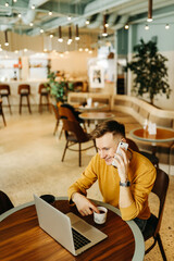 Obraz na płótnie Canvas Young smiling business man freelancer in casual yellow sweater and jeans working remotely using laptop and phone sitting in cafe, selective focus