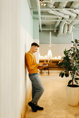 Young smiling business man freelancer in casual yellow sweater and jeans working remotely using laptop and phone sitting in cafe, selective focus