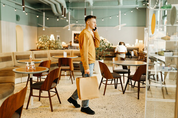 A young man is talking on the phone and carrying paper bags from a store to a cafe