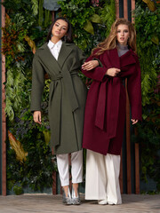 Two girls wearing colorful wool coats and posing against flowal wall. Blonde female model and brunette young woman with long hair standing. Fashion portrait