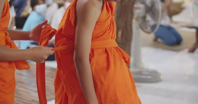 Slow motion of a Buddhist novice who has just passed ordination in the summer during school holidays is helping other novice to dress in a yellow-orange robe.