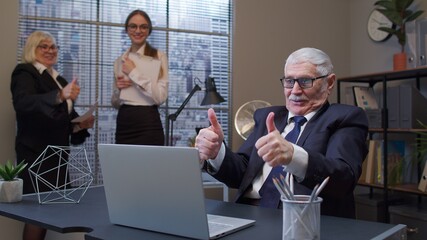 Mature old businessman raising hands celebrating sudden victory with colleagues in office. Cheerful senior business company manager working on portable laptop sitting at desk. Coworking, cooperation