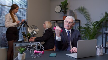 Portrait of smiling business man making fitness exercises with dumbbells after hard work while sits at office desk workplace. Senior old confident manager web designer relaxing. Coworking, freelance