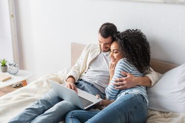 cheerful interracial couple chilling on bed and watching comedy movie on laptop in bedroom