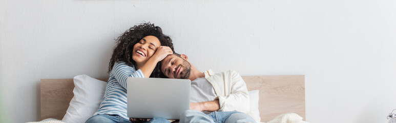 cheerful interracial couple resting in bed near laptop, banner - Powered by Adobe