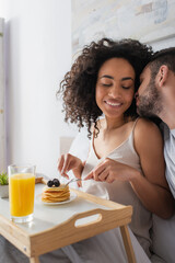 bearded man kissing happy african american woman holding cutlery near pancakes in bedroom