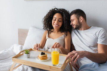 Obraz na płótnie Canvas happy african american woman holding cutlery near pancakes and boyfriend in bedroom