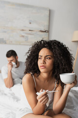 sick african american woman holding digital thermometer and cup near boyfriend on blurred background