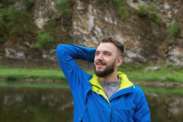 smiling bearded young man looking up at natural background of river and rocky shore. Satisfied with life or successful.