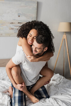 happy bearded man piggybacking african american girlfriend in bedroom
