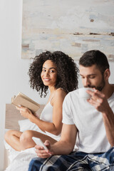 african american woman holding book and looking at boyfriend holding smartphone while drinking coffee on blurred foreground