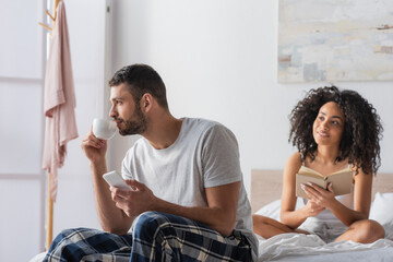 bearded man holding smartphone while drinking coffee and sitting on bed near african american girlfriend reading book o blurred background
