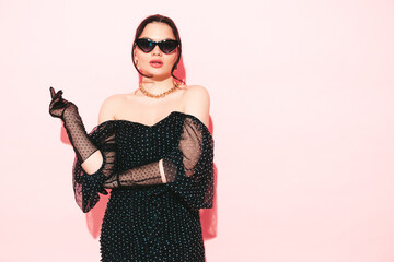 High fashion portrait of young beautiful brunette woman wearing nice trendy summer polka-dot dress and gloves.Sexy fashion model posing near light pink wall in studio.Fashionable female in sunglasses