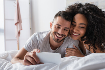 happy multiethnic couple looking at smartphone in bedroom
