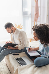 african american woman writing in notebook and looking at laptop near bearded boyfriend holding folder