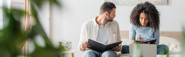 bearded man holding folder with taxes and looking at african american girlfriend with notebook...