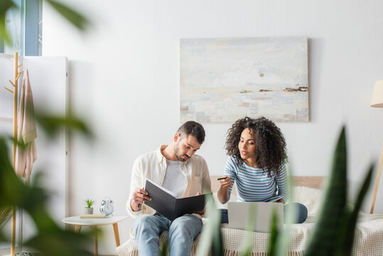 african american woman pointing at folder with taxes near boyfriend and laptop on bed