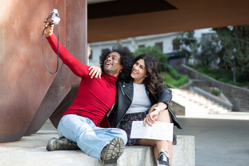 Young multiethnic couple taking smiling photo together with analog camera. They are outdoors. Space for text.