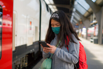 Asian backpacker girl traveling in times of covid19 - young happy and beautiful Korean woman in face mask and backpack checking mobile phone at train station