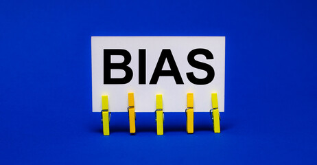 On a bright blue background on yellow clothespins, a white card with the text BIAS