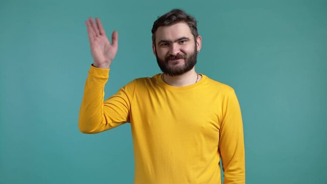 Young friendly man waving hand - hello. Greeting, say Hi to camera. Guy with beard on blue studio background.