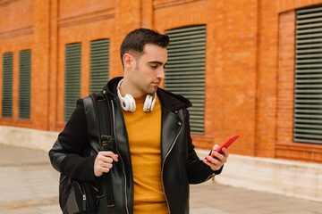 Caucasian young man in yellow sweater and leather jacket using his phone on the street while listening to music and drinking coffee