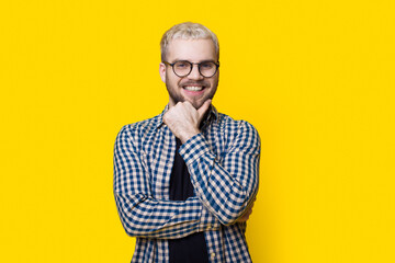 Caucasian man is touching his bearded chin smiling at camera on a yellow studio wall wearing glasses and shirt